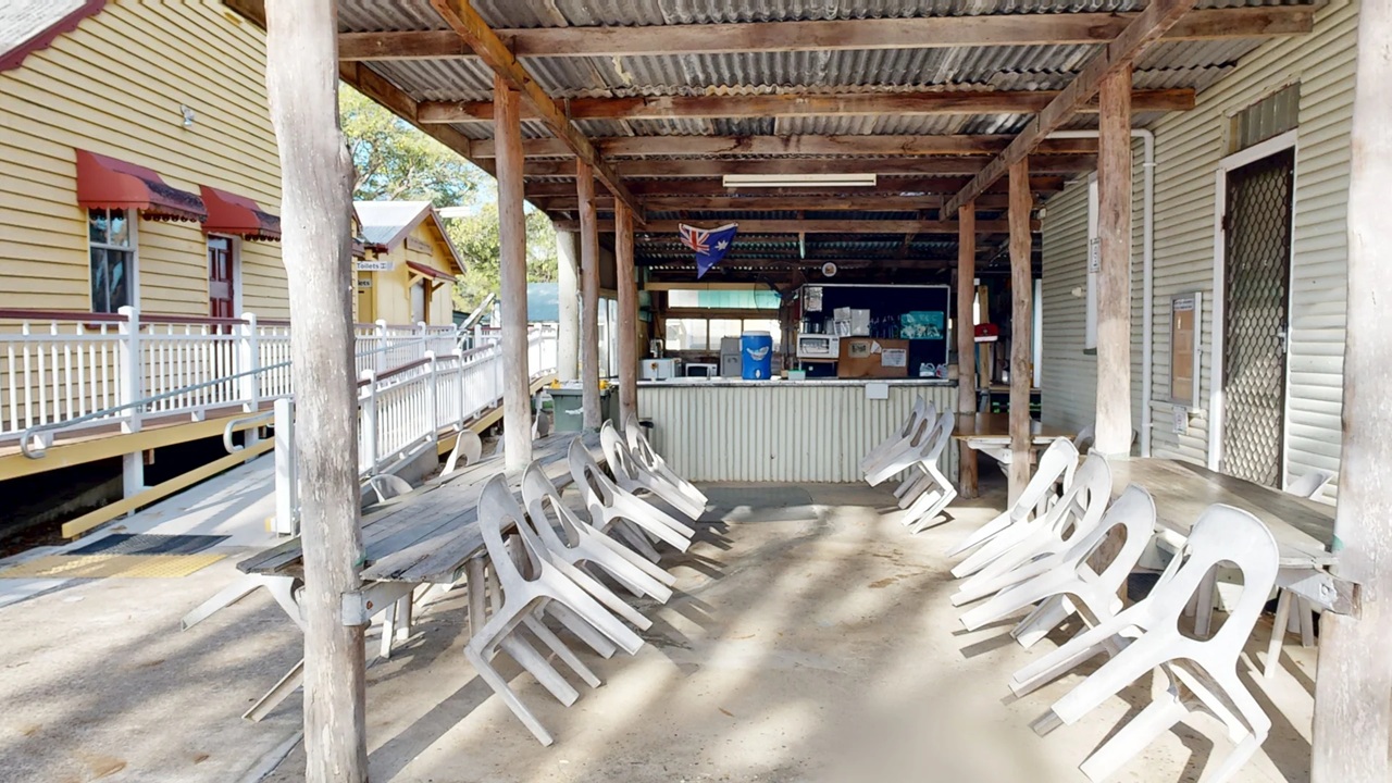 A covered outdoor area with a corrugated metal roof supported by wooden beams. In the centre, a bar or serving counter is visible, with various items like a water cooler and microwave. Plastic chairs are stacked on both sides, leaning against wooden tables. To the left, a wooden building with red awnings and a white railing is visible. The floor is concrete, and the ceiling is made of corrugated metal. Trees can be seen in the background.