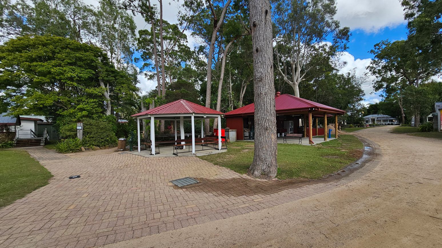 A park area with two red-roofed gazebos on the left, surrounded by tall trees. The foreground has a paved path leading to a dirt road on the right. The gazebos have picnic tables underneath. A large tree trunk is prominent in the centre. In the background, there are more trees and a few small buildings. The grass is neatly trimmed around the structures.