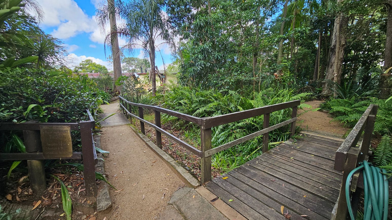 A wooden pathway with railings runs through a lush garden. On the left, a dirt path is bordered by dense green foliage and a small plaque is attached to the railing. On the right, a wooden bridge leads into a forested area with tall trees and ferns. In the background, a yellow building is partially visible among the trees. A garden hose is coiled on the right side of the bridge. The sky is partly cloudy.