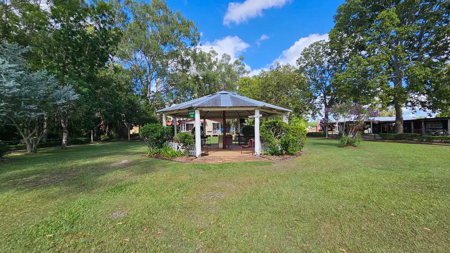 A hexagonal gazebo with a metal roof is centred on a grassy area. It is surrounded by neatly trimmed bushes and small plants. To the left, there are tall trees with dense foliage, while on the right, more trees and a flowering bush are visible. In the background, a building with a veranda can be seen. The sky is blue with scattered clouds.