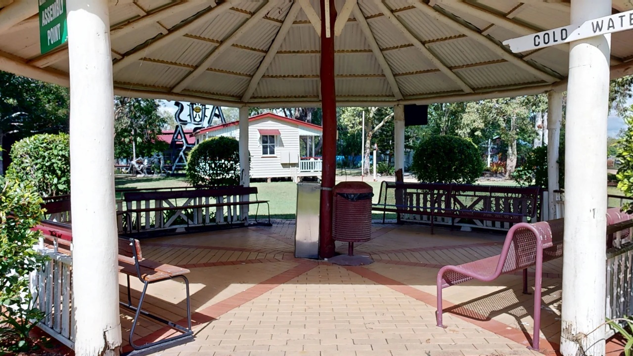 A gazebo with a hexagonal roof supported by white pillars. Inside, there are benches and a red rubbish bin in the centre. The floor is paved with light brown tiles. In the background, a small white building with a red roof is visible, surrounded by greenery and trees. A sign on the right reads "COLD WATER," and another on the left says "ASSEMBLY POINT." Bushes are placed around the perimeter of the gazebo.