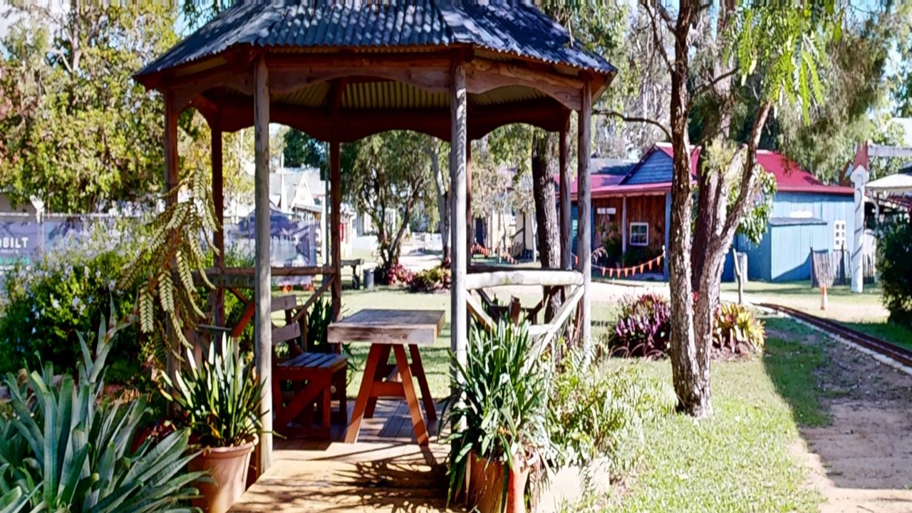A wooden gazebo with a corrugated metal roof is in the centre, surrounded by potted plants and greenery. Inside, there are wooden benches and a table. To the right, a grassy area leads to a path, with trees and a building with a red roof in the background. On the left, more trees and a fence are visible. The scene is well-lit, suggesting a sunny day.
