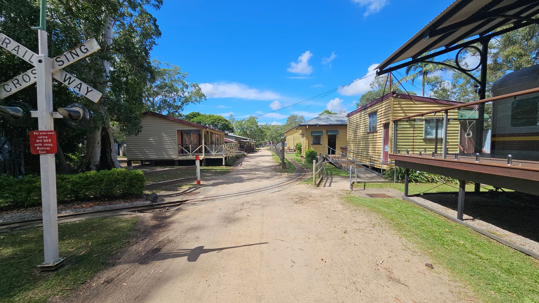 A dirt pathway runs through the centre, flanked by small wooden buildings on both sides. On the left, a railway crossing sign stands near a building with a porch. On the right, a yellow building with a ramp and a glass railing is visible. The path is lined with grass, and trees are scattered throughout the background. A railway track crosses the path, and a red sign warns against walking along the railway line.