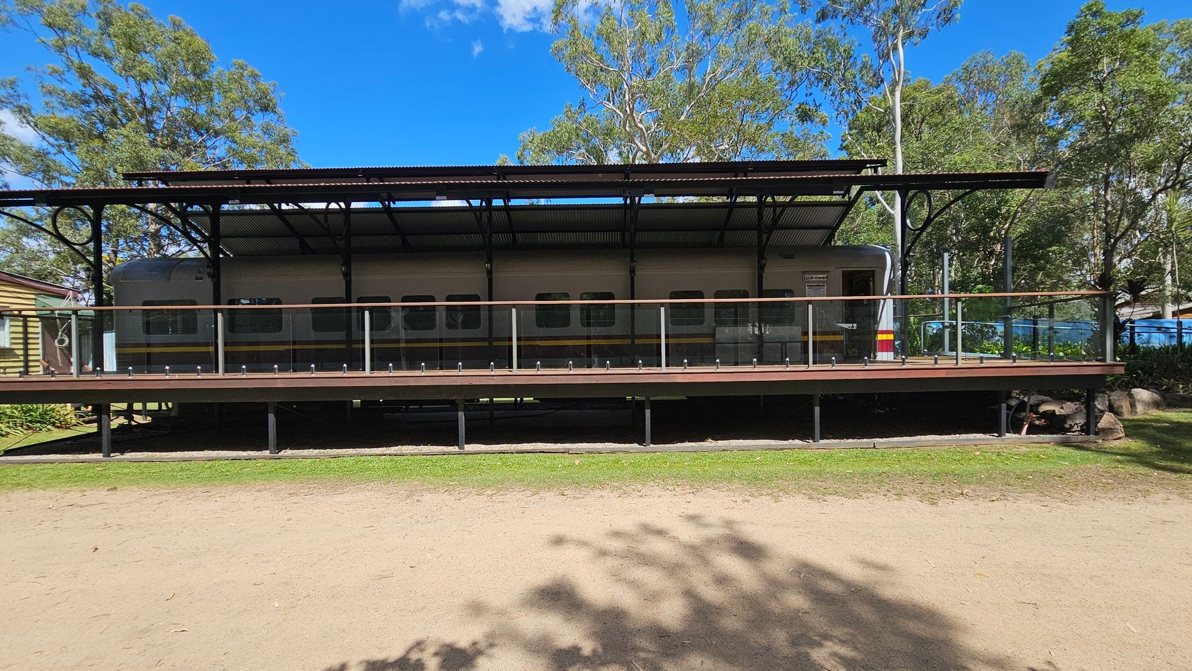 A train carriage is positioned on a raised platform with a metal roof and glass railing. The carriage is grey with a yellow and red stripe. In the background, there are tall trees with green foliage. The ground in the foreground is a mix of grass and dirt, with some shadows cast by the trees. On the left, part of a wooden building is visible.