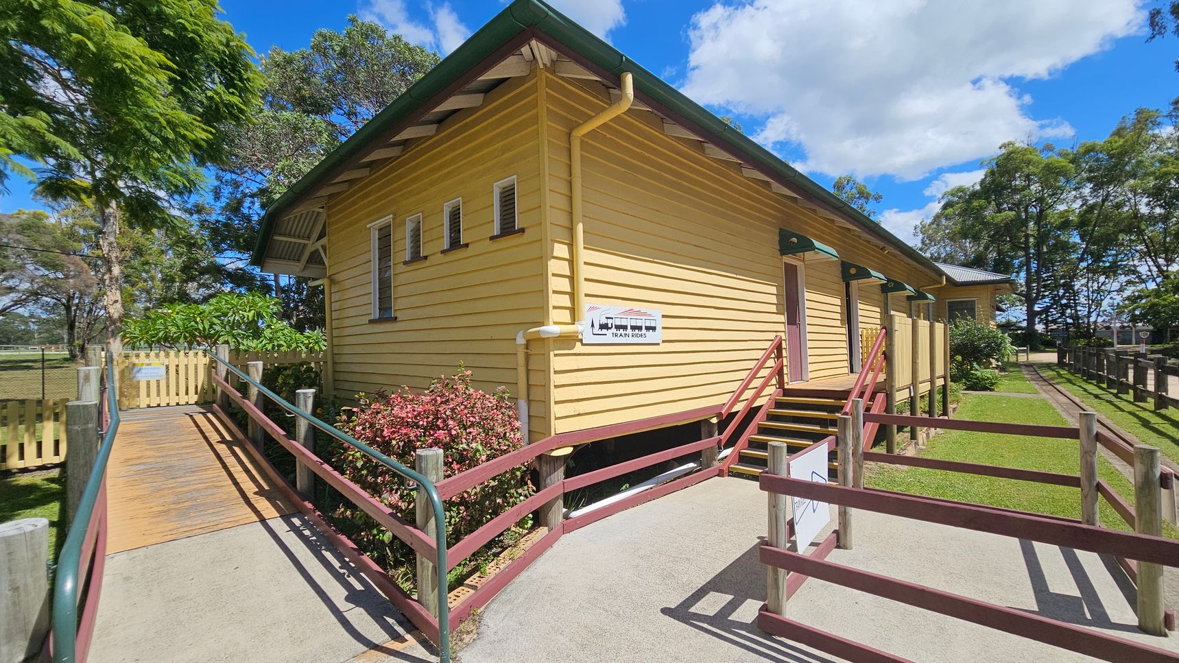 A yellow wooden building with a green roof and a sign indicating "Train Rides" on the side. A wooden ramp with green railings leads to the entrance on the left, surrounded by trees and a small garden with pink flowers. On the right, there are steps with a handrail leading to another entrance. The ground is a mix of concrete and grass, and a wooden fence lines the path. The sky is partly cloudy.