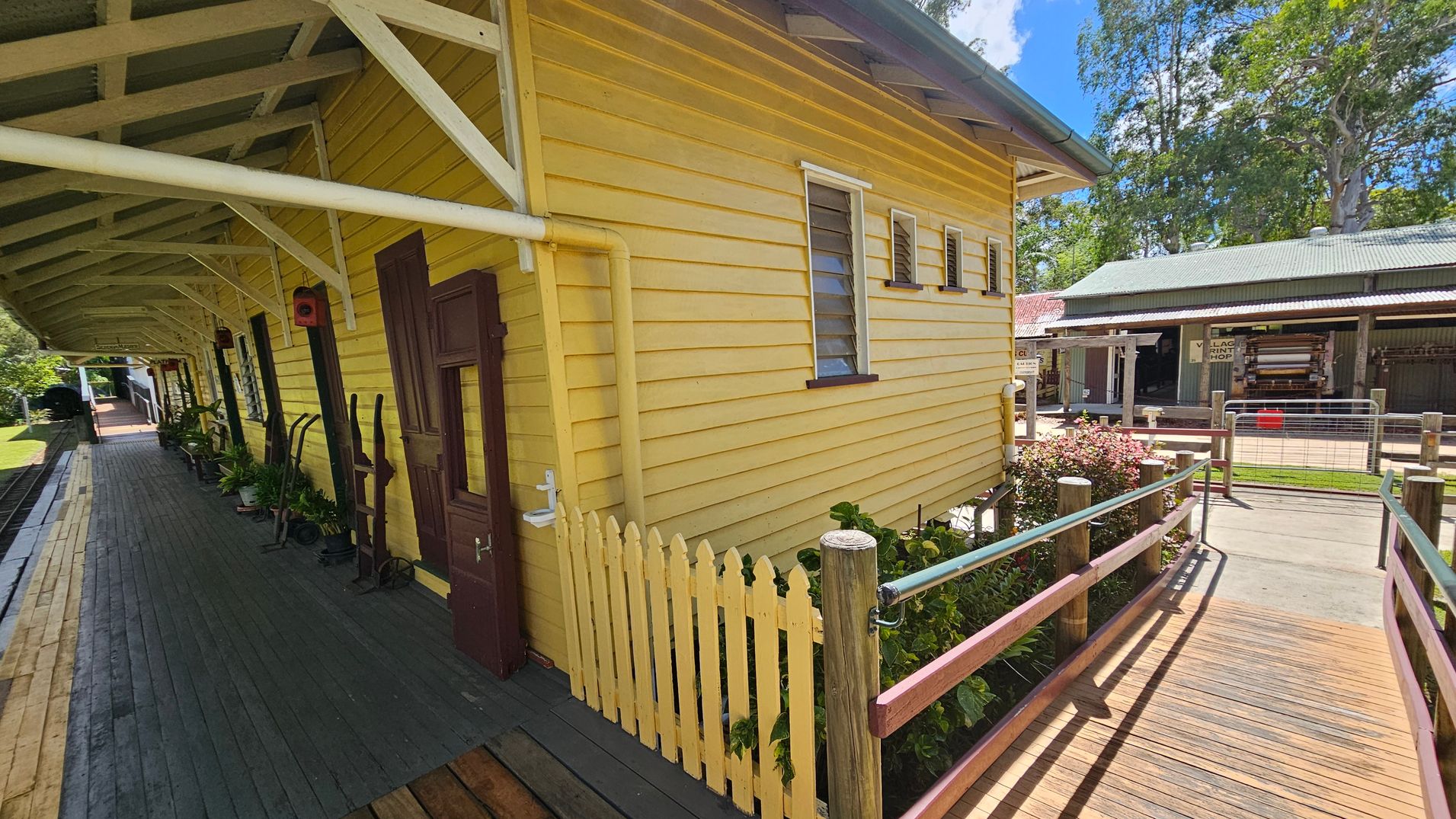 A yellow wooden building with a sloped roof and several small windows on the right. A wooden ramp with a railing leads up to the building, bordered by a small yellow picket fence and plants. On the left, a covered wooden platform with trolleys and potted plants runs alongside the building. In the background, another structure with a corrugated metal roof and open sides is visible, surrounded by trees. The ground is paved with concrete.