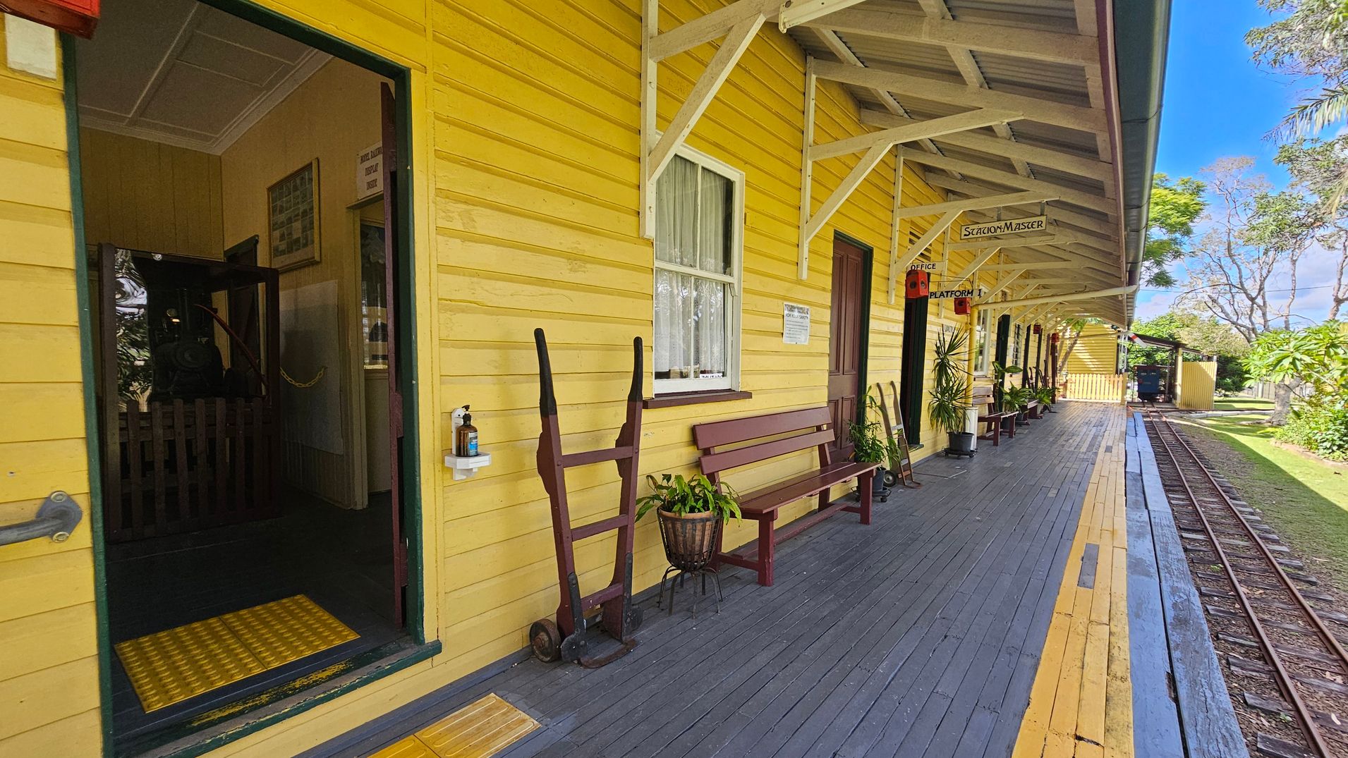 A vintage railway station platform with a yellow wooden building on the left. The building has a green-trimmed door and window, with a bench and a hand trolley nearby. A hand sanitiser dispenser is mounted on the wall. The platform is wooden with yellow and grey planks, leading to a narrow railway track on the right. Signs reading "Station Master" and "Platform 1" hang from the roof. The background features trees and a grassy area.
