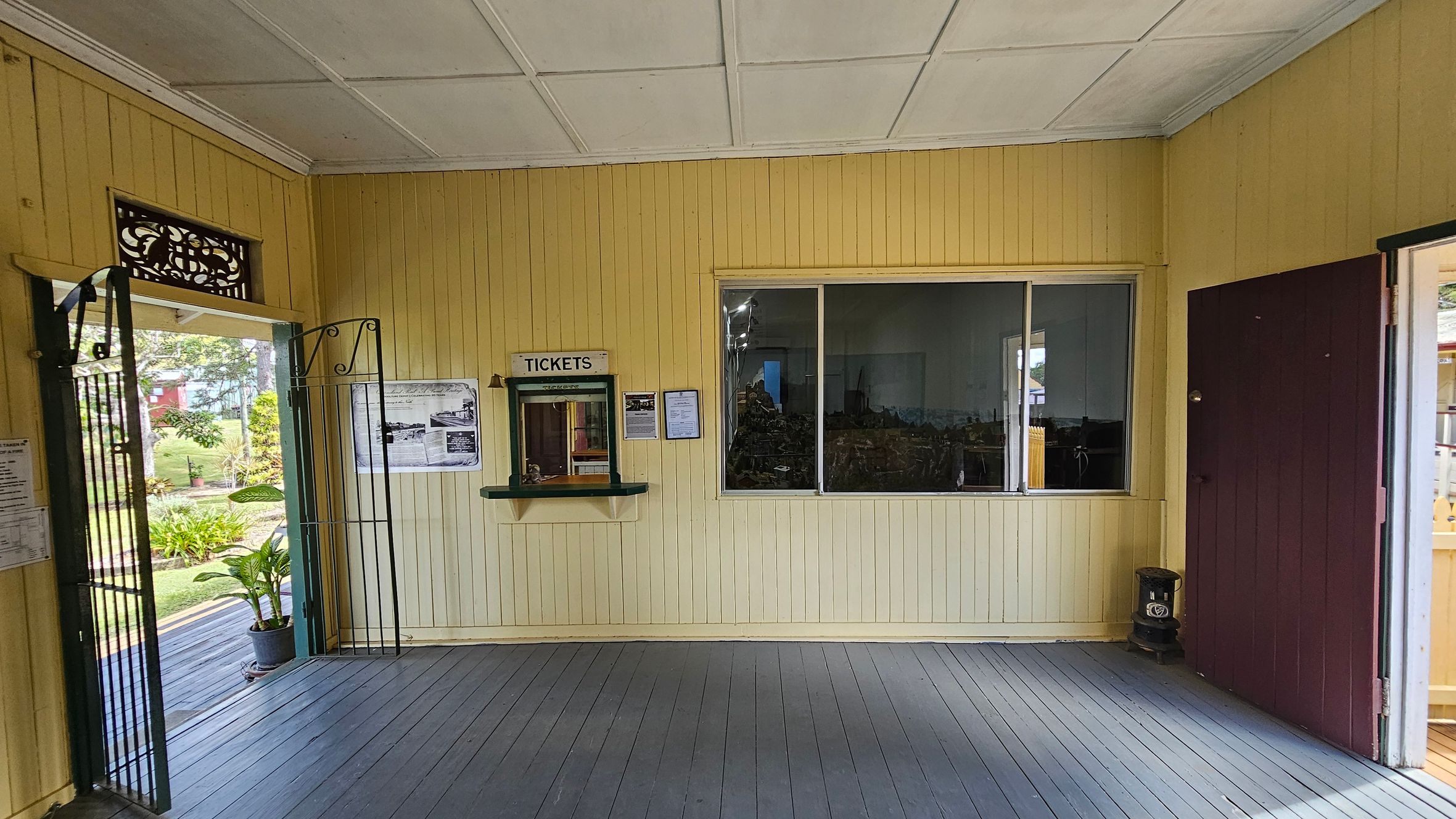 A room with light yellow wooden walls and a grey panelled ceiling. On the left, an open metal gate leads outside, revealing greenery and a potted plant. In the centre, a ticket window with a "TICKETS" sign is flanked by historical photos and information. A large window is next to it. On the right, a closed maroon door. The floor is made of dark grey wooden planks.
