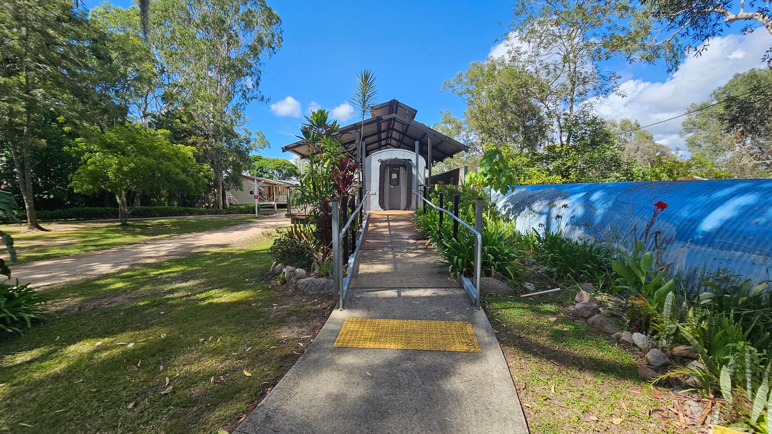 A concrete path with a yellow tactile paving leads to a structure resembling a train carriage, covered by a metal roof. The path is flanked by lush green plants and small trees. On the left, a dirt path and more trees are visible, along with a small building. On the right, a blue corrugated fence runs parallel to the path, with more greenery and a single red flower. Tall trees surround the area, providing a natural backdrop.