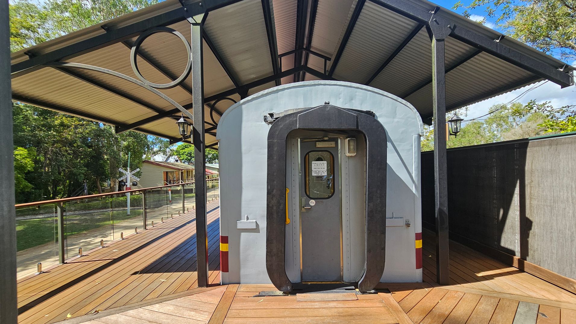 A vintage train carriage is positioned on a wooden platform with a metal roof overhead. The carriage door is central, featuring a small window and a sign. To the left, a glass railing runs along the platform, with trees and a building visible in the background. A railway crossing sign is also present. The platform is made of wooden planks, and the roof is supported by black metal beams with decorative circular elements.