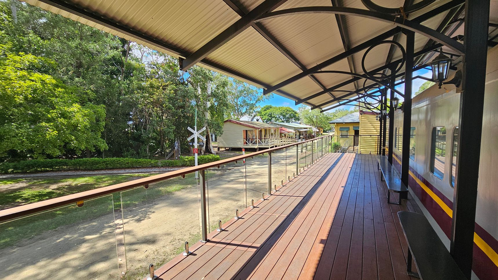 A wooden platform with a metal railing runs alongside a train carriage on the right. The platform is covered by a roof with black metal supports. Benches are placed along the carriage. On the left, there is a grassy area with dense green trees. In the background, several small wooden buildings are visible, along with a railway crossing sign. The ground is a mix of grass and dirt.