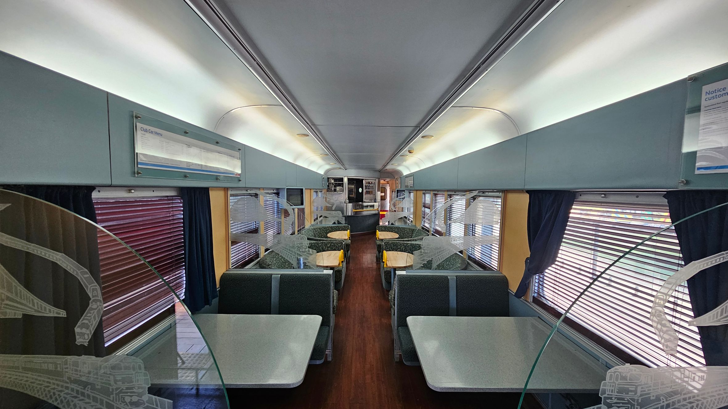 Interior of a train dining car with a central aisle. On both sides, there are booth-style seating areas with grey tables and patterned green seats. The windows have horizontal blinds and dark curtains. Above the windows, there are light blue panels with menus and notices. The ceiling is white with recessed lighting. At the far end, there is a service counter with shelves and a doorway. The floor is wooden, and decorative glass panels with train motifs separate the seating areas.