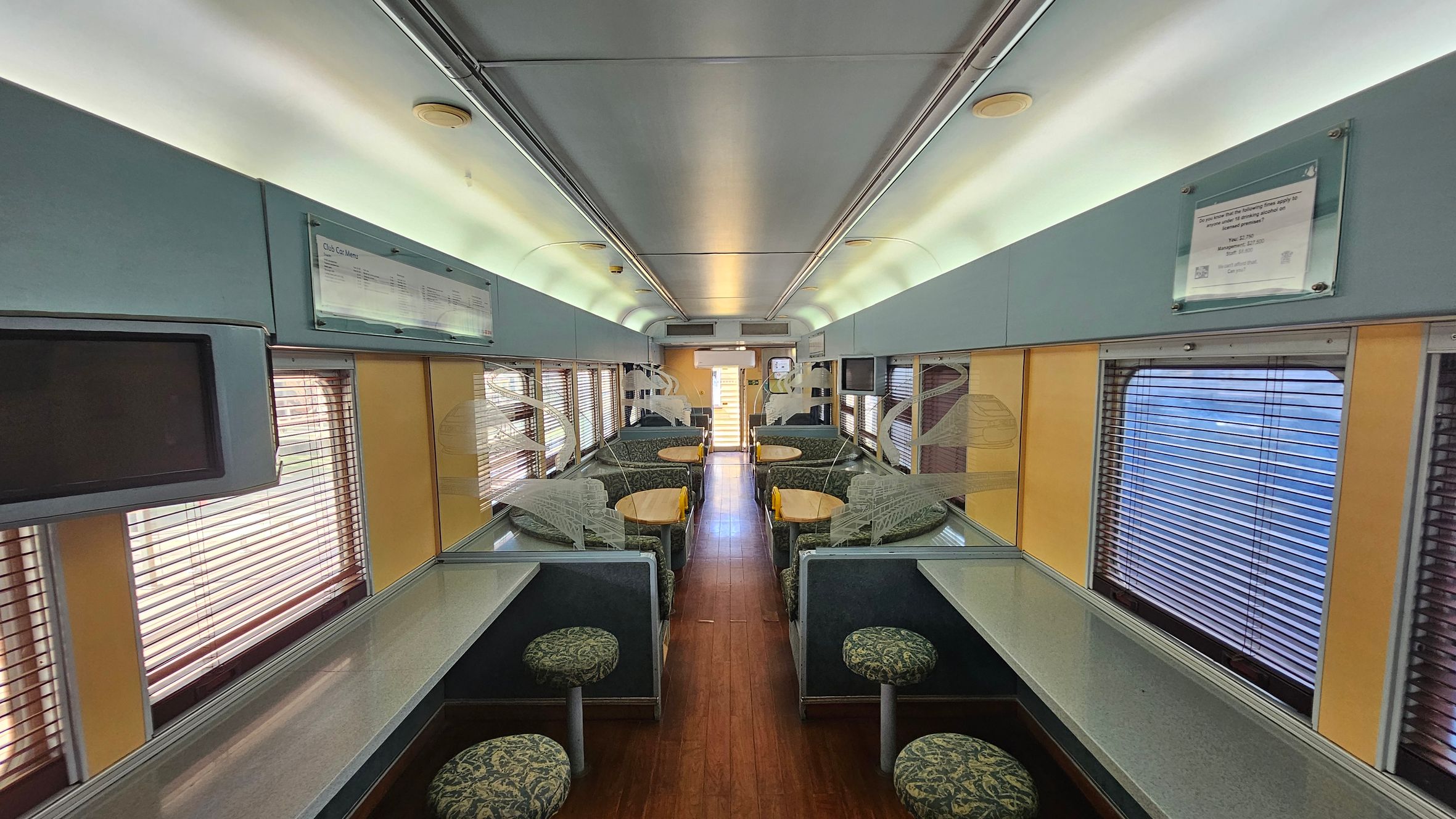 Inside a train carriage with a central aisle flanked by seating areas. On both sides, there are tables with green patterned stools and cushioned benches. The windows have blinds partially drawn. Overhead, the ceiling is curved with lighting along the edges. The floor is wooden, and there are signs and screens mounted on the walls above the windows. The overall colour scheme includes shades of green and beige.