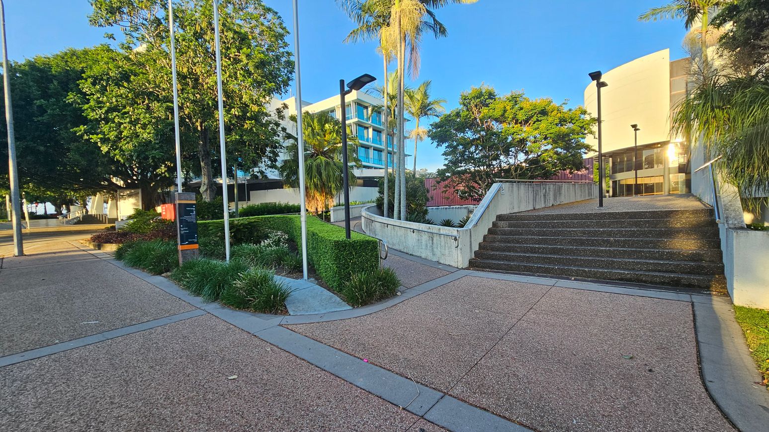 A paved pathway with a fork in the middle, leading to a set of stairs on the right. The stairs are bordered by a low concrete wall with metal railings. On the left, there are flagpoles and a sign surrounded by neatly trimmed hedges and various plants. Tall palm trees and a large leafy tree provide shade. In the background, a modern building with blue-tinted windows is visible. The area is well-lit with street lamps.