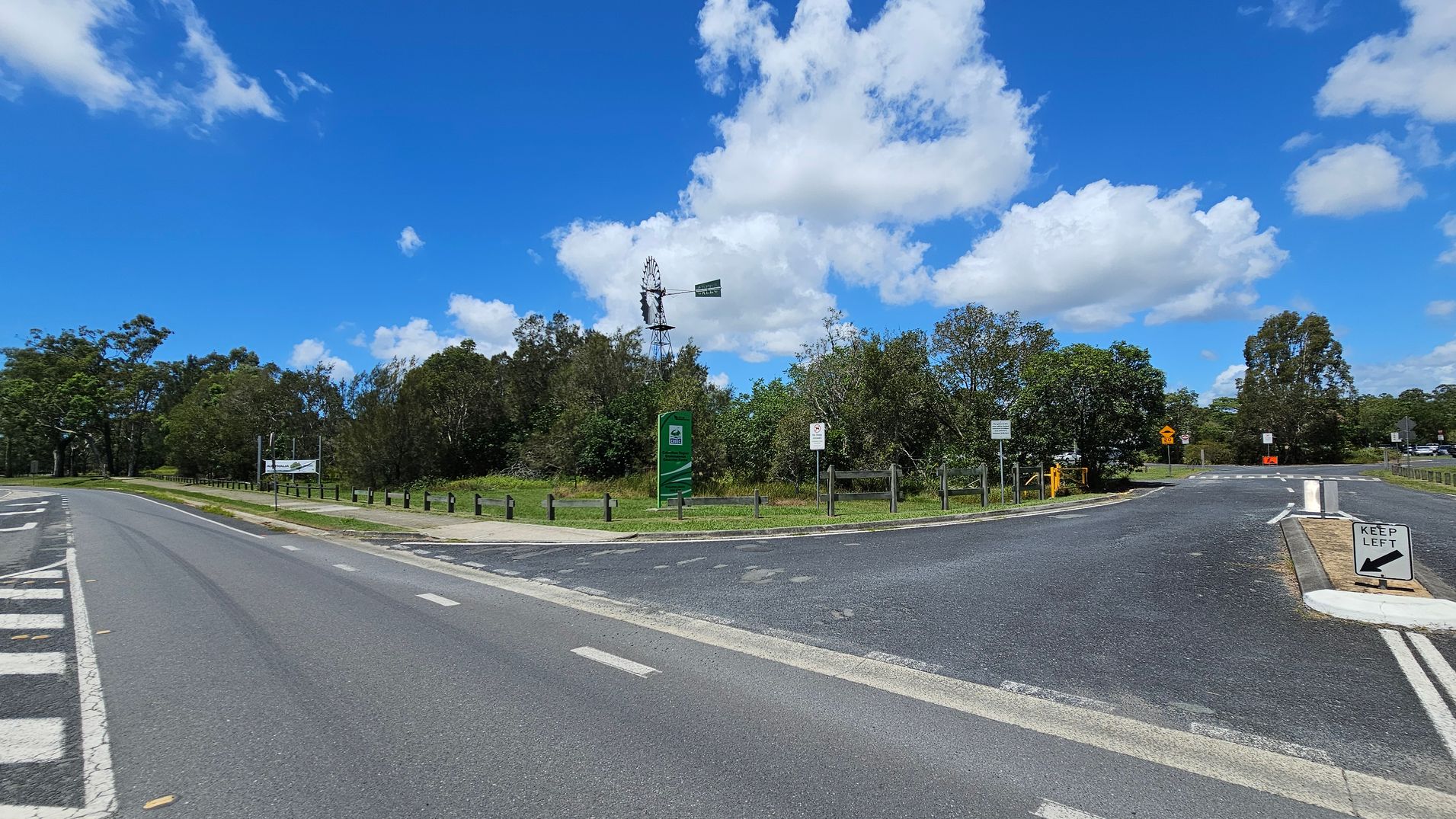 A road with a "Keep Left" sign in the foreground. On the left, a road curves with a line of wooden posts and a sign. In the centre, a green sign and a windmill are visible, surrounded by trees and bushes. On the right, another road with a small traffic island. The background is filled with more trees under a blue sky with scattered clouds.