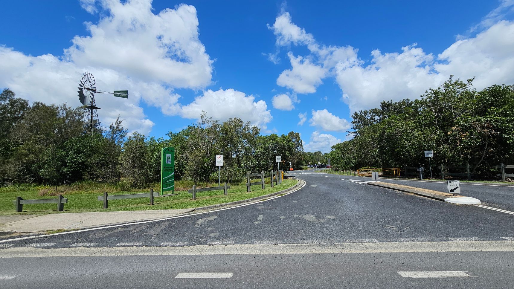 A road curves to the right, bordered by green grass and trees. On the left, a tall windmill stands among the trees, with a green sign nearby. Several smaller signs are visible along the road, including a "Keep Left" sign on a small island. The road surface shows some wear, and wooden barriers line the grassy areas. The sky is blue with scattered clouds.