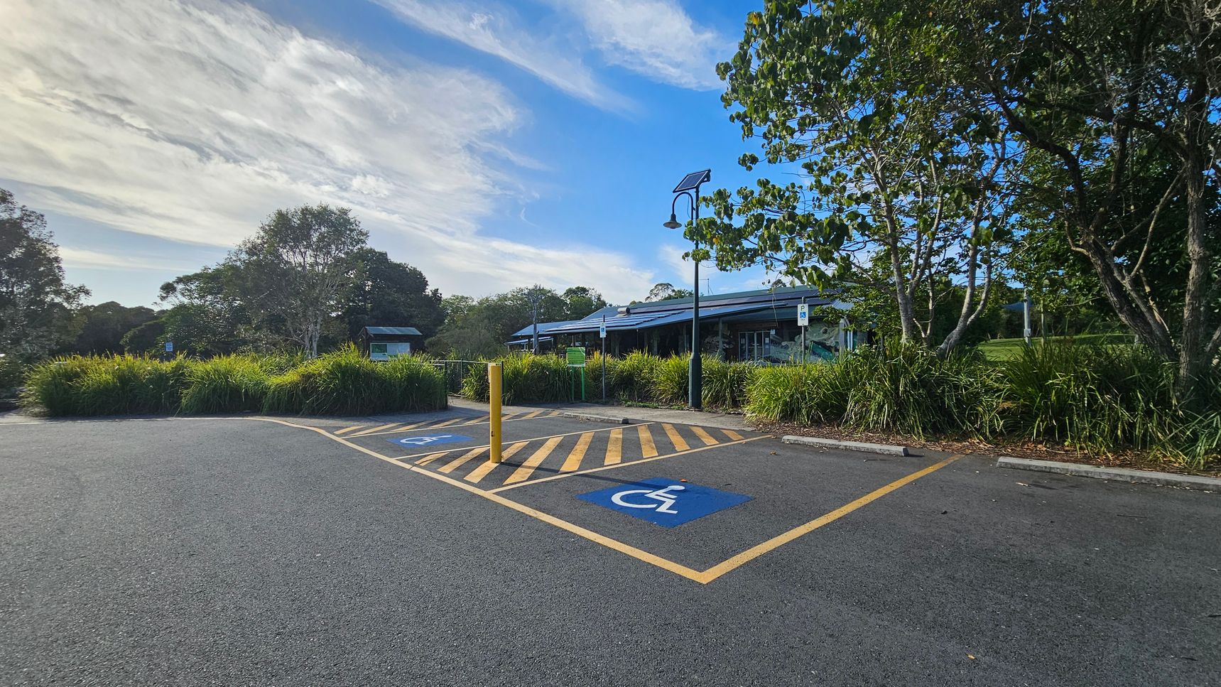A car park with two marked disabled parking spaces in the foreground, featuring blue wheelchair symbols. Yellow diagonal lines separate the spaces. A yellow bollard stands between them. In the background, there is a building with a sloped roof, partially obscured by trees and bushes. A lamp post with a solar panel is visible near the building. The area is surrounded by greenery, including tall grasses and trees.