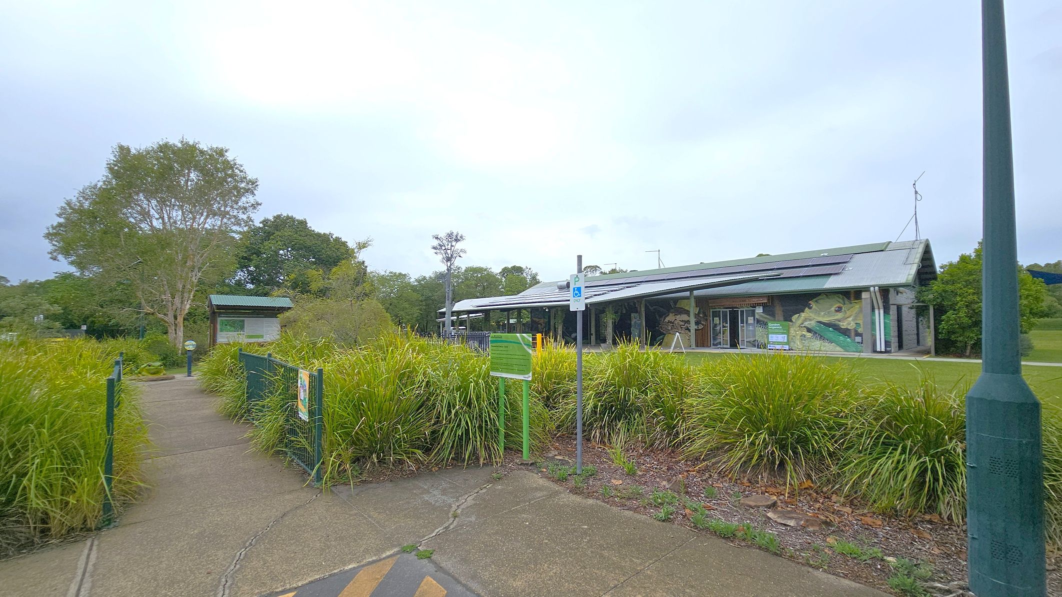 A pathway leads through lush greenery towards a building with a sloped roof on the right. The building features a mural and solar panels. A signpost with a parking sign is visible near the path. On the left, a smaller structure is partially obscured by trees and bushes. The ground is a mix of concrete and grass, and a tall lamppost stands in the foreground.