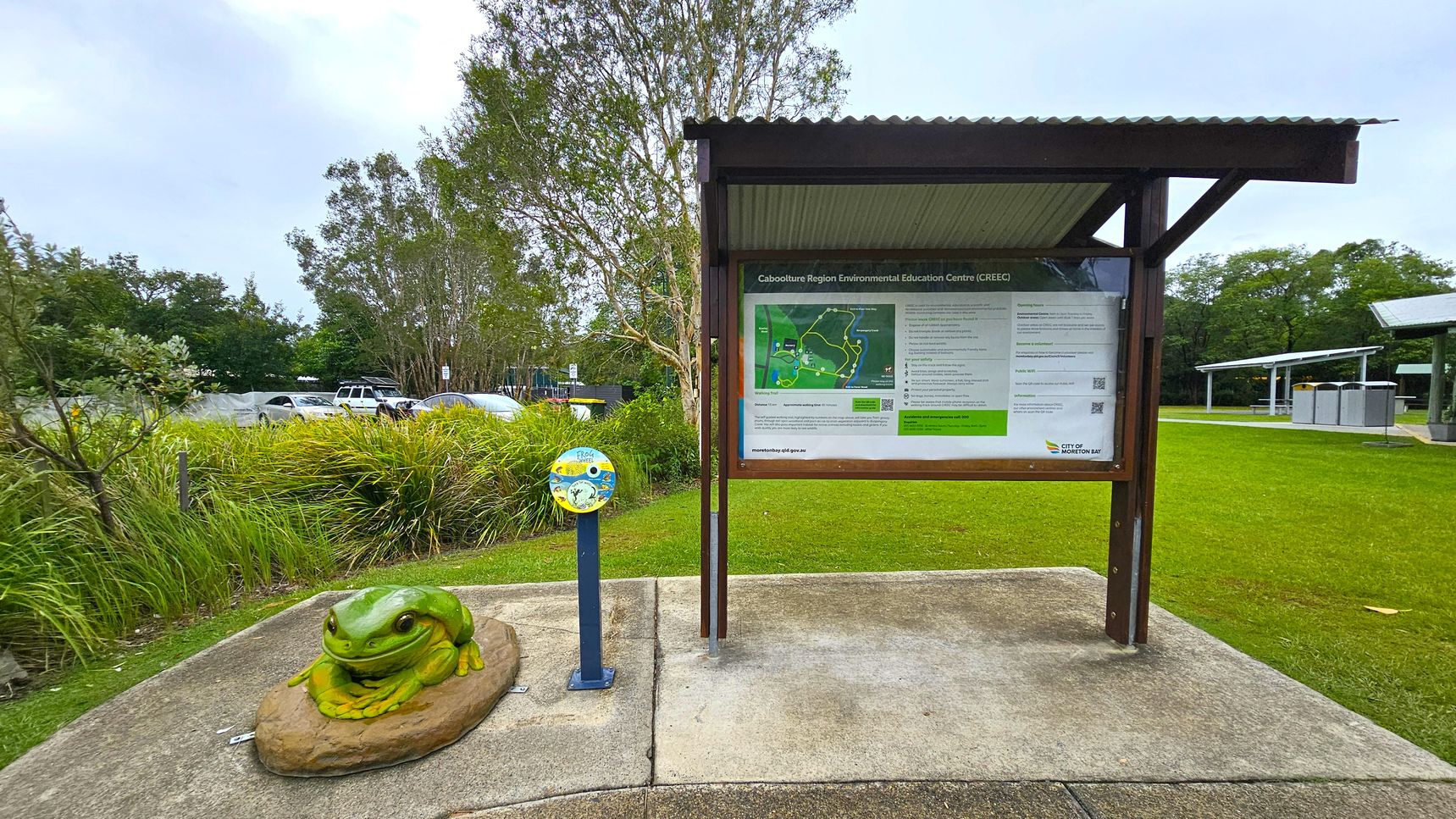 A green frog sculpture sits on a rock to the left, surrounded by grass and plants. In the centre, a signboard under a corrugated metal roof displays information about the Caboolture Region Environmental Education Centre. To the right, a grassy area extends with a few trees and a structure with solar panels. In the background, parked cars are partially visible behind the vegetation. The ground is paved with concrete.