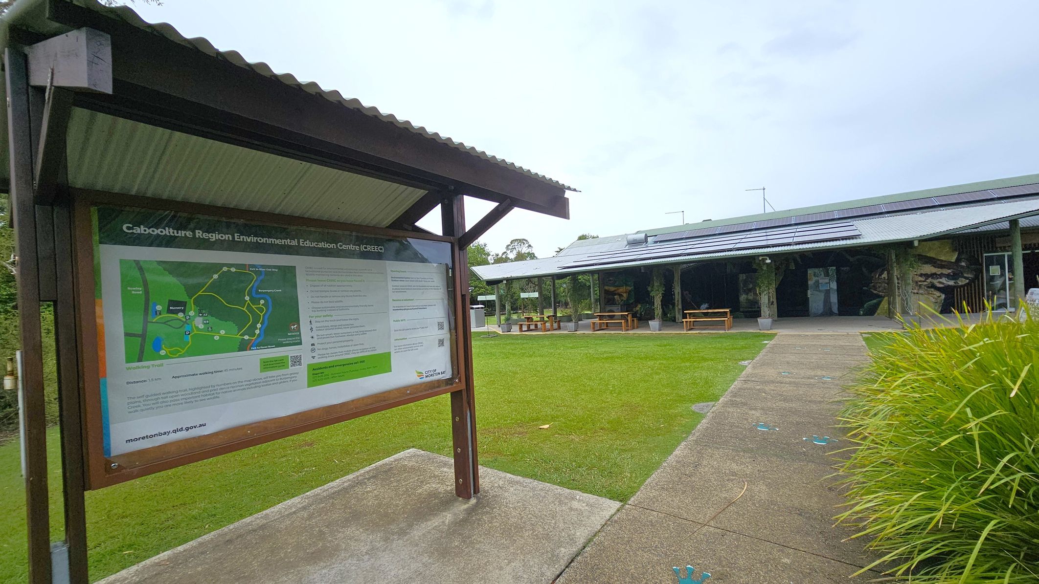 A covered information board on the left displays a map and details about the Caboolture Region Environmental Education Centre. The board is supported by wooden posts. On the right, a building with a corrugated metal roof and solar panels is visible, featuring open sides and picnic tables. The foreground has a paved path leading to the building, bordered by green grass and plants.
