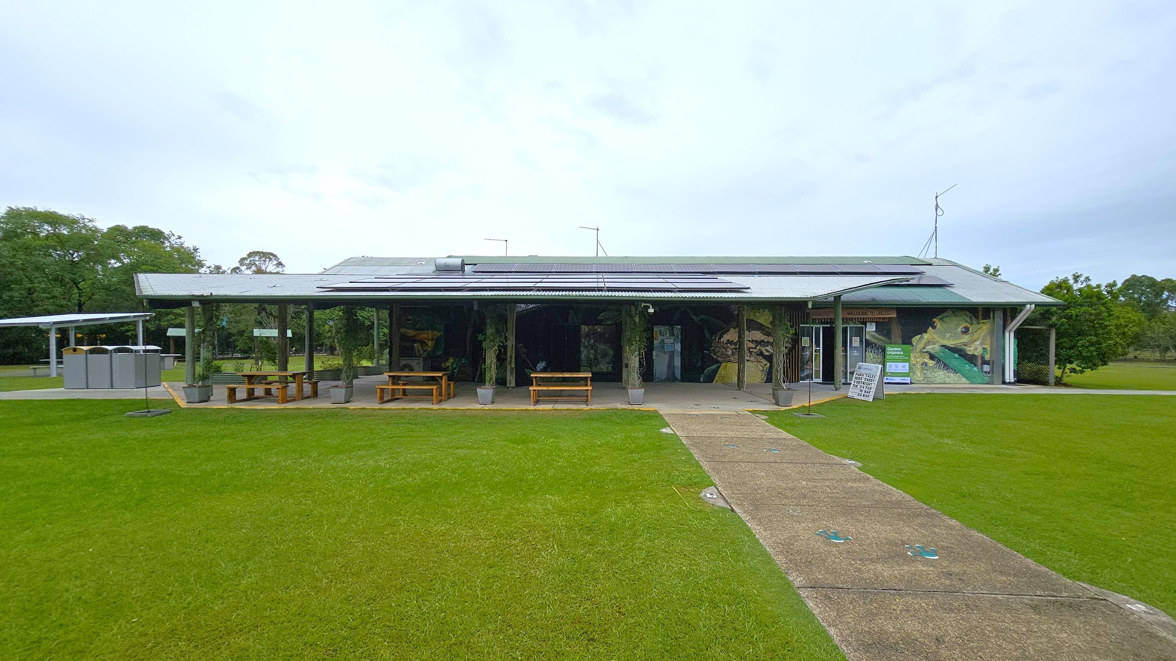A single-storey building with a corrugated metal roof and solar panels. The front features a covered area with wooden picnic tables and potted plants. A mural of a large frog is painted on the right side. A signboard is near the entrance. The foreground has a grassy lawn with a concrete path leading to the building. On the left, there are several portable toilets. Trees and greenery are visible in the background.