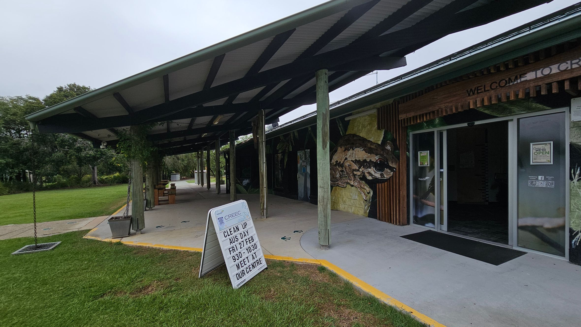 A building with a sloped roof supported by wooden posts. On the right, there's a glass entrance door with a sign above it. A large mural of a frog is painted on the wall next to the door. In the foreground, a sandwich board sign on the grass displays event information. The left side features a grassy area with trees in the background. The floor is concrete, and the ceiling is made of corrugated metal.