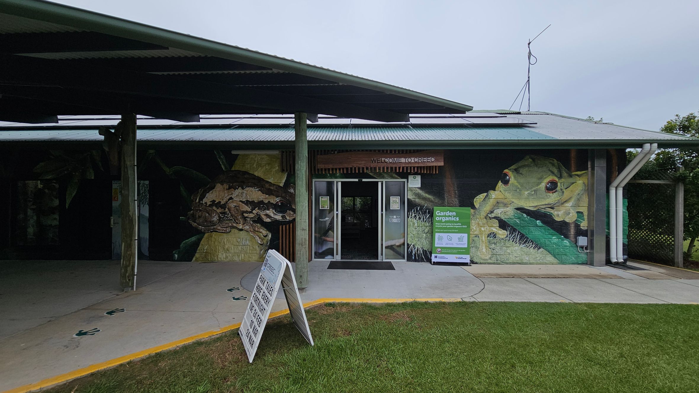 A building with a corrugated metal roof and a mural of large frogs on the front wall. The entrance has glass doors with a "Welcome to GREEC" sign above. To the right of the entrance, there's a sign about garden organics. A sandwich board sign is on the pavement in front, and the ground is a mix of concrete and grass. The ceiling is not visible.