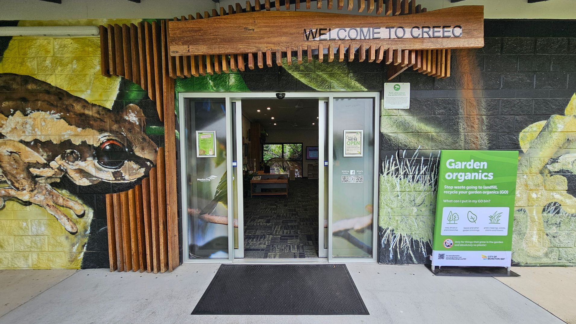 A building entrance with glass sliding doors is in the centre, flanked by a mural of a frog on the left wall. Above the doors, a wooden sign reads "WELCOME TO CREEC". To the right of the entrance, a green sign titled "Garden organics" provides recycling information. The floor is concrete with a black mat in front of the doors. Inside, a room with a patterned carpet and a window at the back is visible.