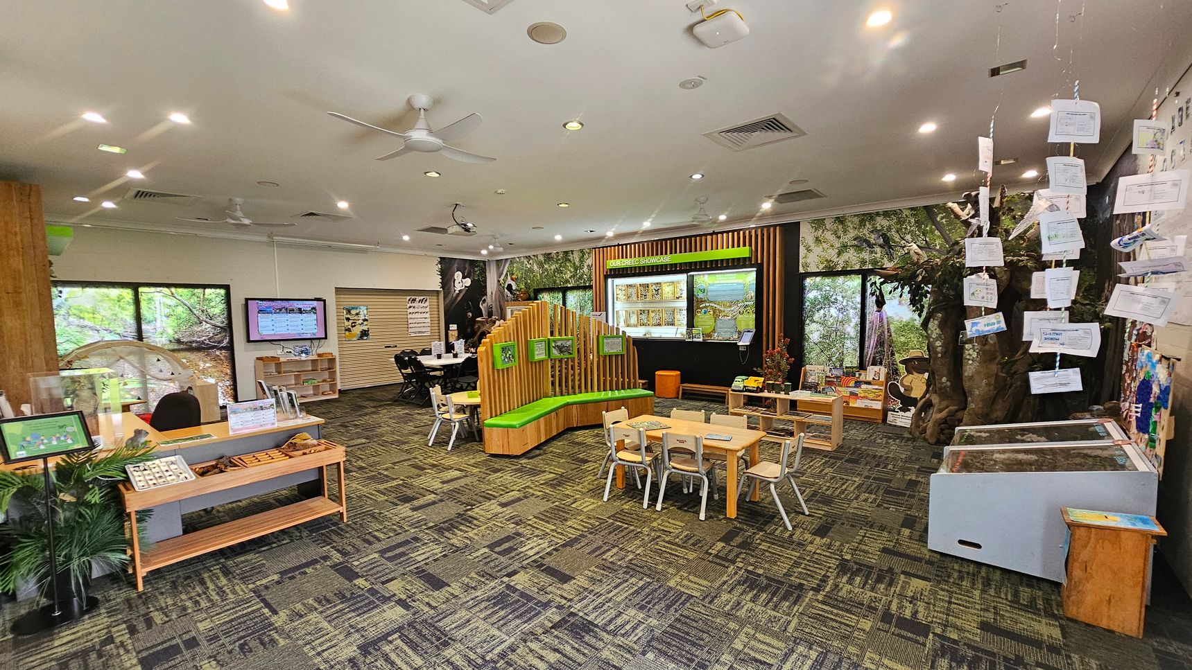 A spacious room with a patterned carpet floor and a ceiling with recessed lighting and ceiling fans. On the left, there is a wooden desk with a display and a small shelf. In the centre, a curved wooden bench with green cushions is surrounded by small tables and chairs. The right side features a tree-like structure with hanging papers and a display case. The back wall has large windows with nature scenes and a display area labelled "Our Creek Showcase."