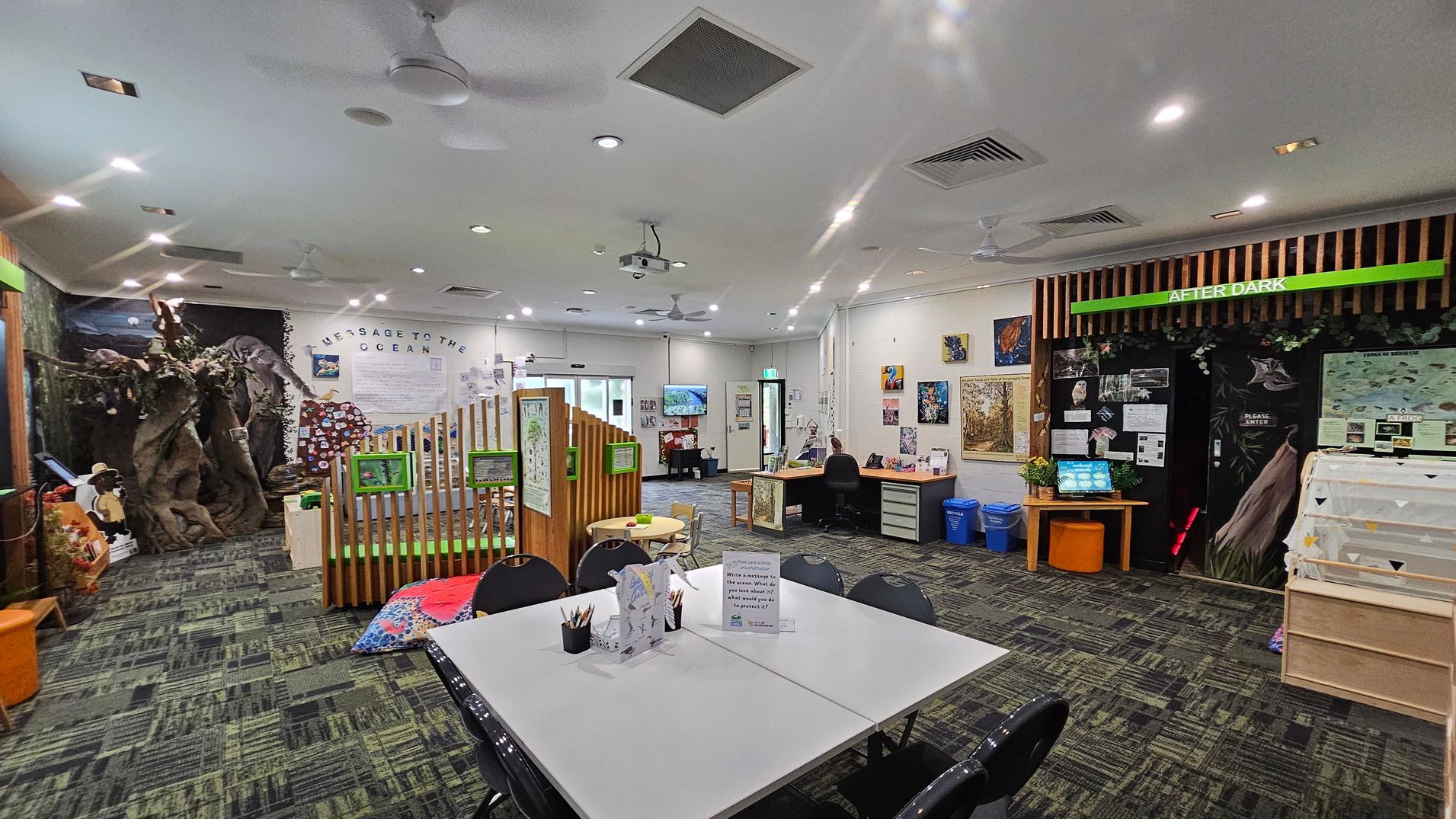 A spacious room with a green and grey patterned carpet. On the left, there's a large tree structure and a wooden fence with green signs. In the centre, a table with chairs and art supplies. The back wall has a "Message to the Ocean" display. On the right, a section labelled "After Dark" with a computer and informational posters. The ceiling is white with recessed lighting and ceiling fans. Walls are adorned with various artworks and educational materials.