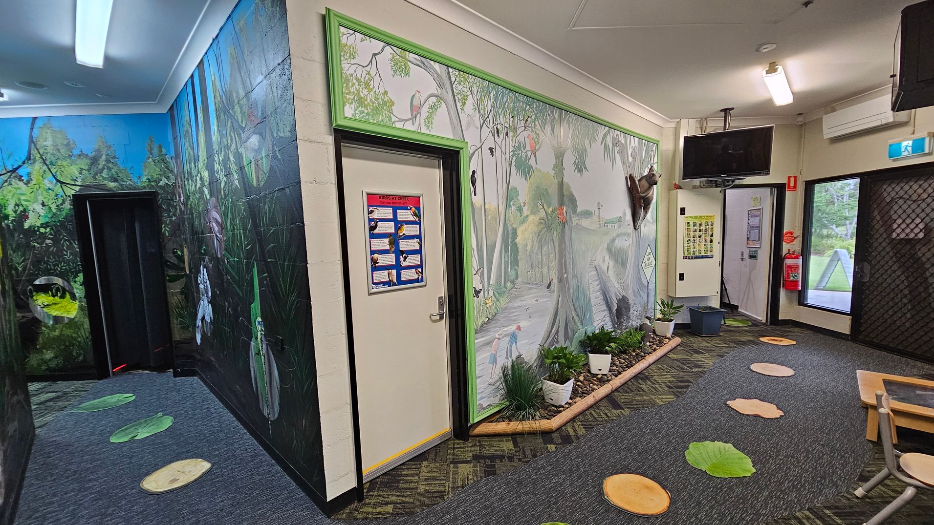 A hallway with vibrant wall murals depicting a forest scene. On the left, a dark doorway is surrounded by lush greenery and wildlife illustrations. The right wall features a mural with trees and birds, bordered by a green frame. A door with a poster is in the centre. The floor has a patterned carpet with leaf and log designs. Ceiling lights illuminate the space, and a TV is mounted near the ceiling. A fire extinguisher and exit sign are visible near a glass door.