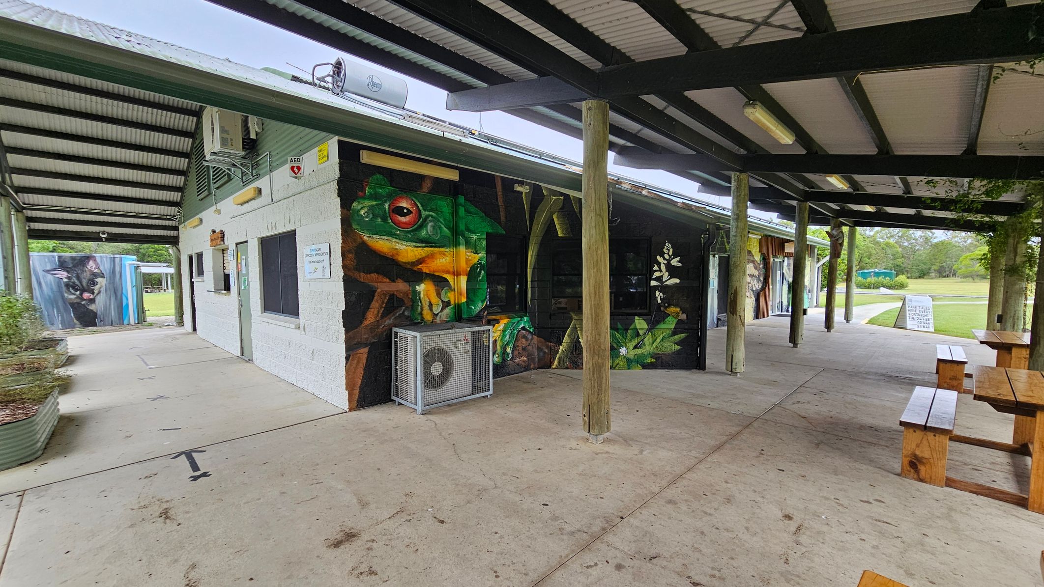 A building with a corrugated metal roof and a mural of a large green and yellow frog on the wall. To the left, there's a mural of a cat on a separate structure. The building has an air conditioning unit mounted on the wall. Wooden picnic tables are on the right under a covered area supported by wooden posts. The floor is concrete, and the background features a grassy area with trees. A sign is visible near the grass.
