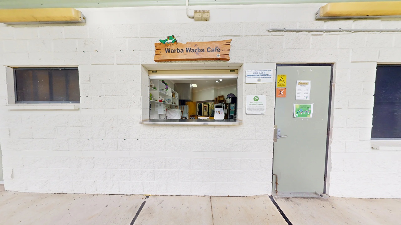 A small café window set in a white brick wall with a wooden sign above reading "Warba Warba Cafe". The window is open, revealing shelves with various items inside. To the right of the window is a grey door with several signs and notices attached. On either side of the window and door are small, dark-tinted windows. The ground is paved with light-coloured concrete slabs.