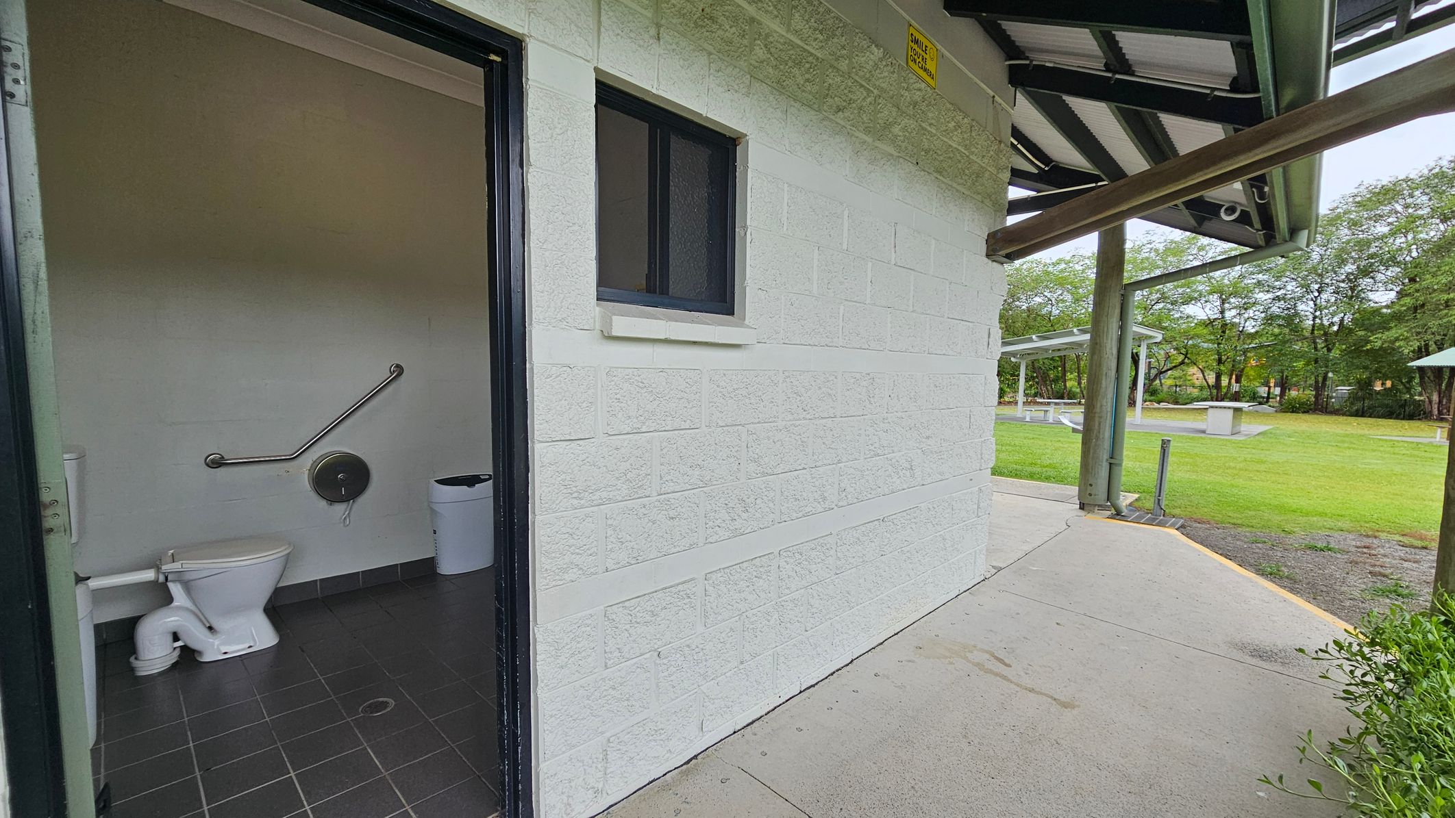 A public toilet with a white brick exterior wall and a small window. The door is open, revealing a toilet with a grab bar and a bin inside. The floor is tiled in dark grey. Outside, there's a concrete path leading to a grassy area with trees and a picnic shelter in the background. The roof has a metal structure supported by wooden beams. A small sign is visible near the roof.