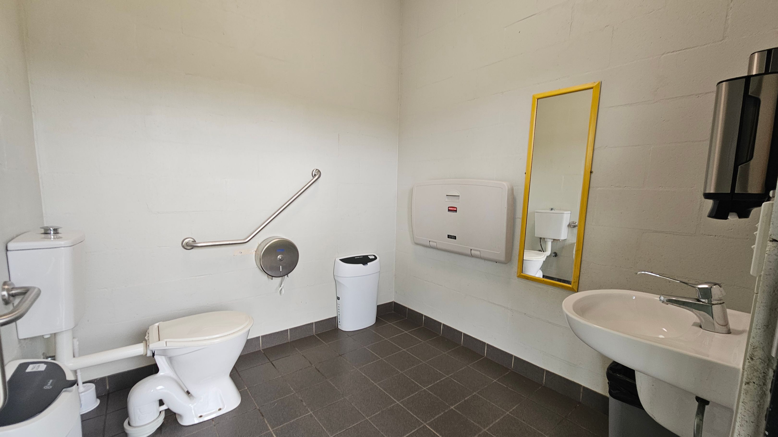 A public restroom with a white toilet on the left, equipped with a grab bar and a toilet paper dispenser. A white bin is positioned nearby. On the right, a white sink with a silver tap is mounted on the wall, accompanied by a soap dispenser. A yellow-framed mirror is above a baby changing station. The floor is tiled in dark grey, and the walls are light-coloured.