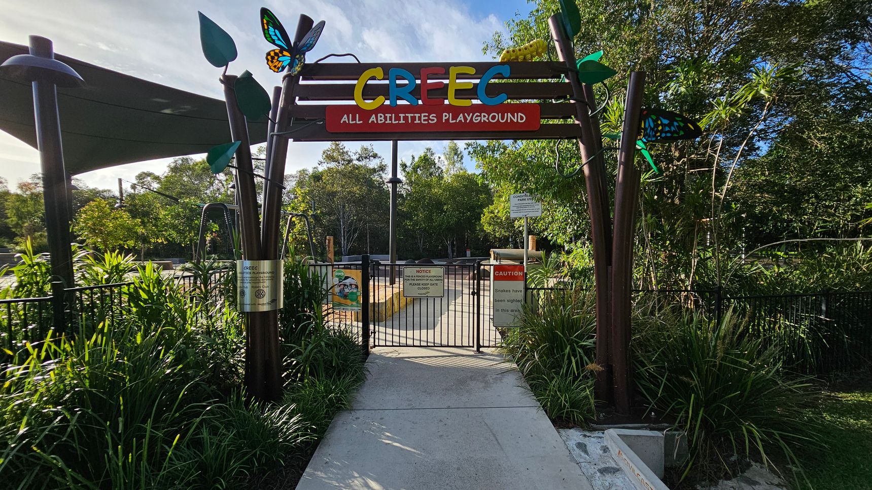 Entrance to a playground with a colourful sign reading "CREEC All Abilities Playground" at the top. The gate is black metal with various signs, including a caution about snakes. Decorative elements like butterflies and leaves adorn the entrance. Lush greenery surrounds the area, with trees and plants on both sides. A shaded area with a canopy is visible on the left. The path leading into the playground is concrete.