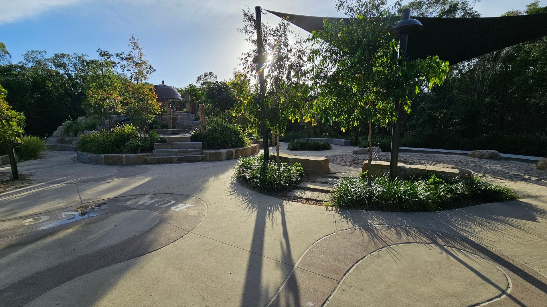 A park setting with a paved pathway curving through the foreground. On the left, stone steps lead up to a small structure surrounded by lush greenery. In the centre, a tree casts long shadows across the ground, with sunlight filtering through its leaves. To the right, a sandy area is partially shaded by a triangular canopy. The background is filled with dense trees and foliage.