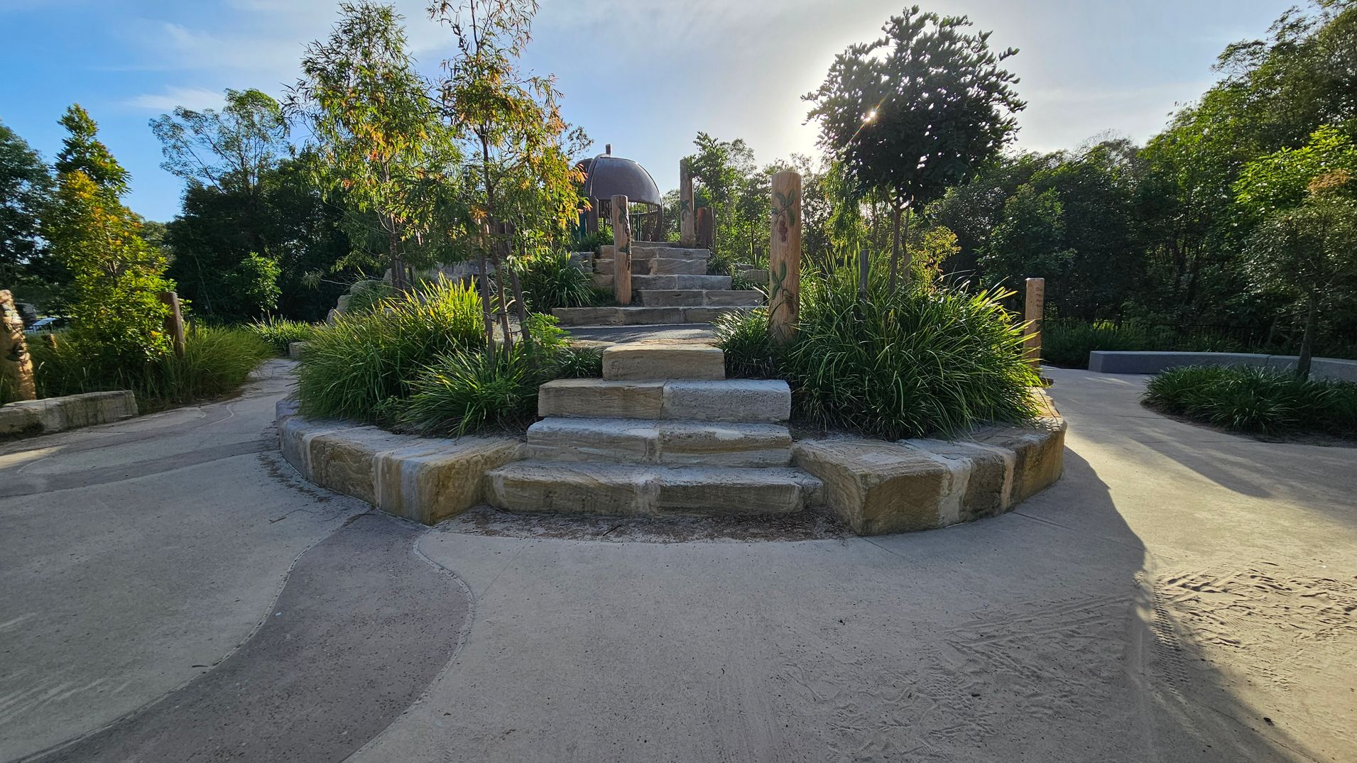 Stone steps lead up to a landscaped area with lush green plants and small trees. The path is bordered by stone walls, and wooden posts are visible on either side. The background is filled with dense trees, and sunlight filters through, casting shadows on the ground. The path is smooth and curves gently around the landscaped area.