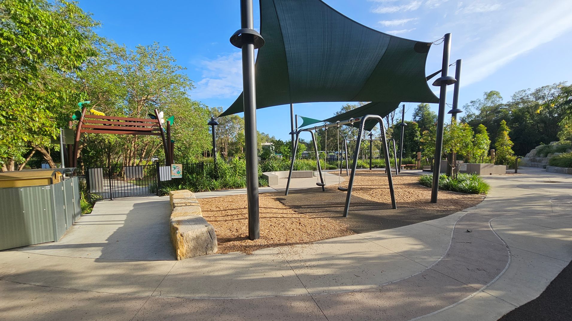 A playground with swings is on the right, shaded by a large dark green canopy. The ground is covered with a mix of concrete and mulch. On the left, a wooden archway with green and yellow decorations leads to a gated area surrounded by lush greenery. In the background, there are more trees and landscaped areas. The path curves gently through the scene, and there are stone benches and planters with small trees and shrubs.