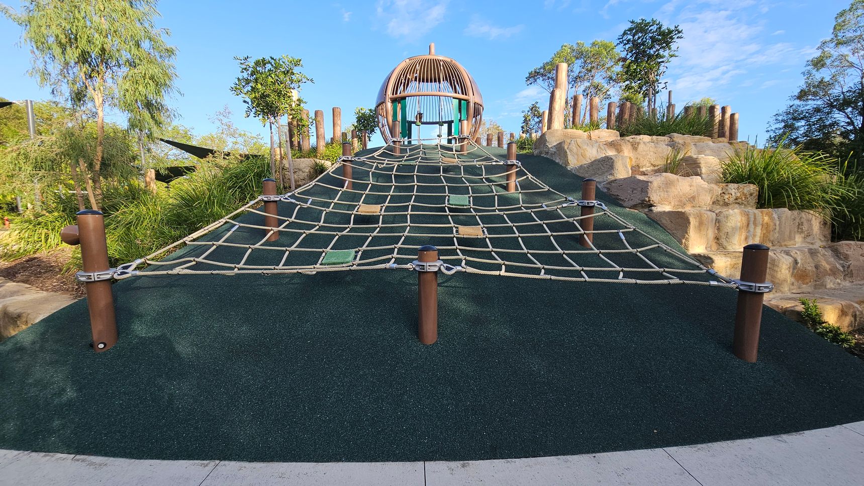 A playground with a large climbing net in the centre, supported by wooden posts. The net leads up to a wooden structure resembling a dome. On the right, there are stone steps with grass and wooden posts. On the left, trees and shrubs are visible. The ground is covered with a dark green rubber surface, and a concrete path runs along the bottom edge.