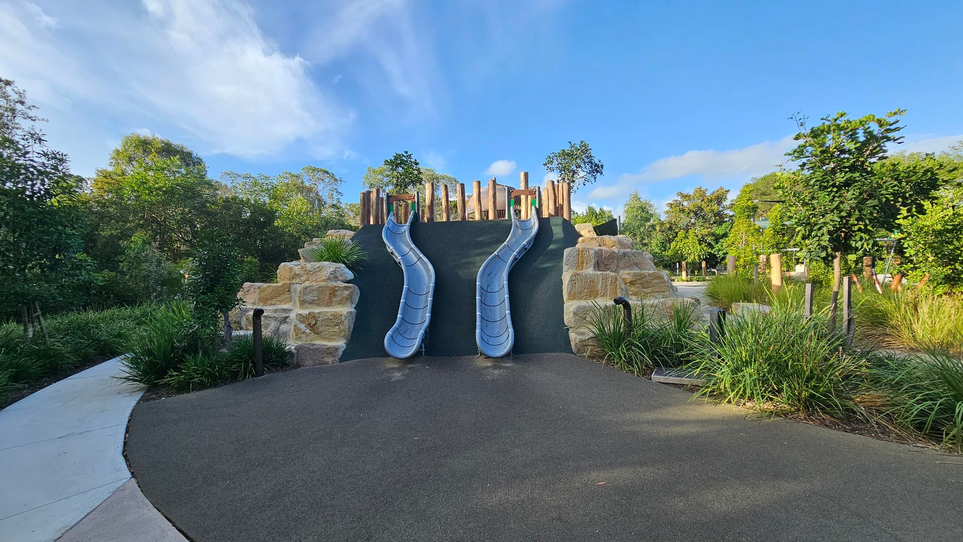 Two blue slides are positioned side by side, descending from a structure made of large stone blocks and wooden posts. The slides are set against a dark surface, with greenery and small trees surrounding the area. The foreground features a smooth, dark pathway leading up to the slides, bordered by grass and plants. The background is filled with lush trees and foliage, creating a natural setting.