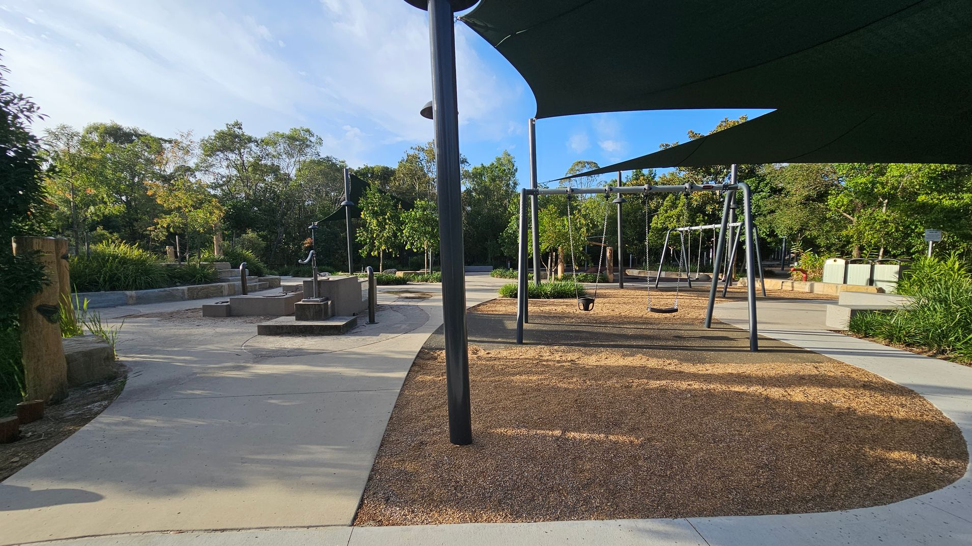 A playground with a swing set on the right, featuring multiple swings, including a toddler seat. The ground is covered with wood chips. On the left, there are water play features with metal pumps and a concrete path winding through. The area is shaded by large, dark green canopies supported by black poles. In the background, there are lush green trees and shrubs, with more pathways and seating areas visible. The sky is clear and blue.