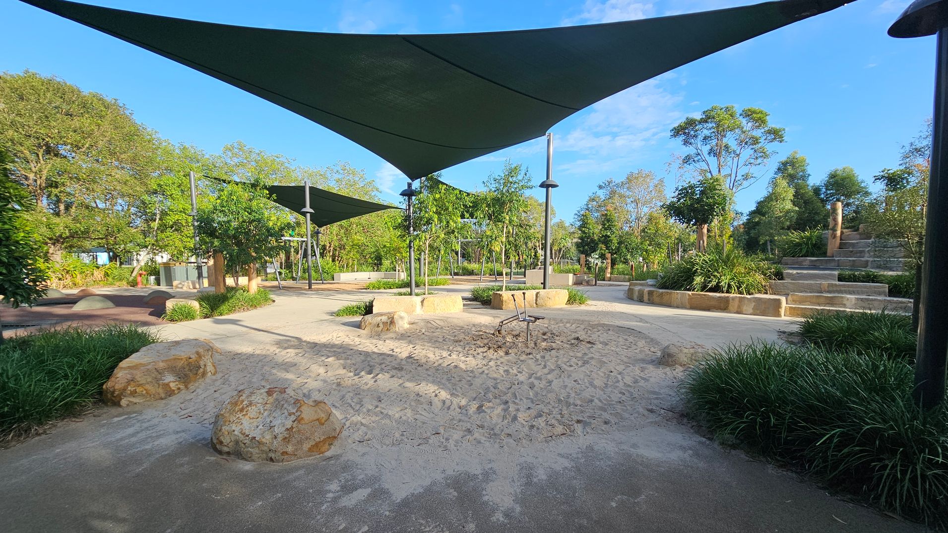 A park with a sandy play area in the centre, surrounded by large rocks. Overhead, there are triangular shade sails supported by metal poles. To the right, stone steps lead up a small hill, bordered by lush greenery. Trees and shrubs are scattered throughout the background, with a clear blue sky above. The ground is a mix of sand and paved pathways, with patches of grass and plants around the edges.