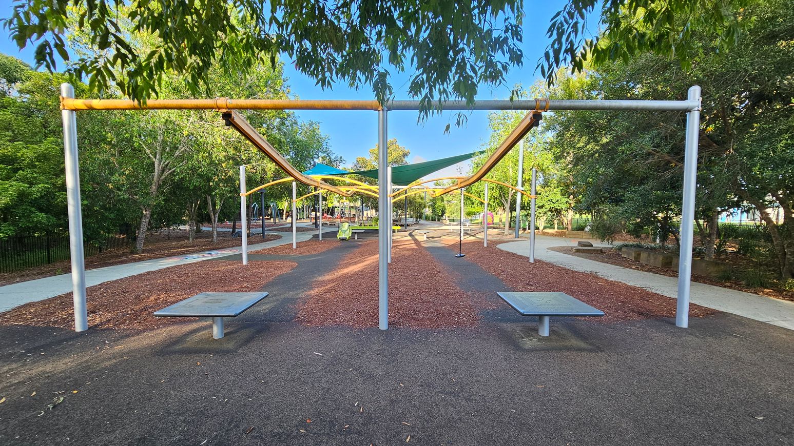 A park scene with a pathway running through the centre, flanked by trees on both sides. Overhead, there are yellow and green shade structures supported by metal poles. The ground is covered with a mix of dark and reddish-brown surfaces. In the foreground, there are two metal benches on either side of the path. The background features more trees and park equipment, with a clear sky visible through the foliage.