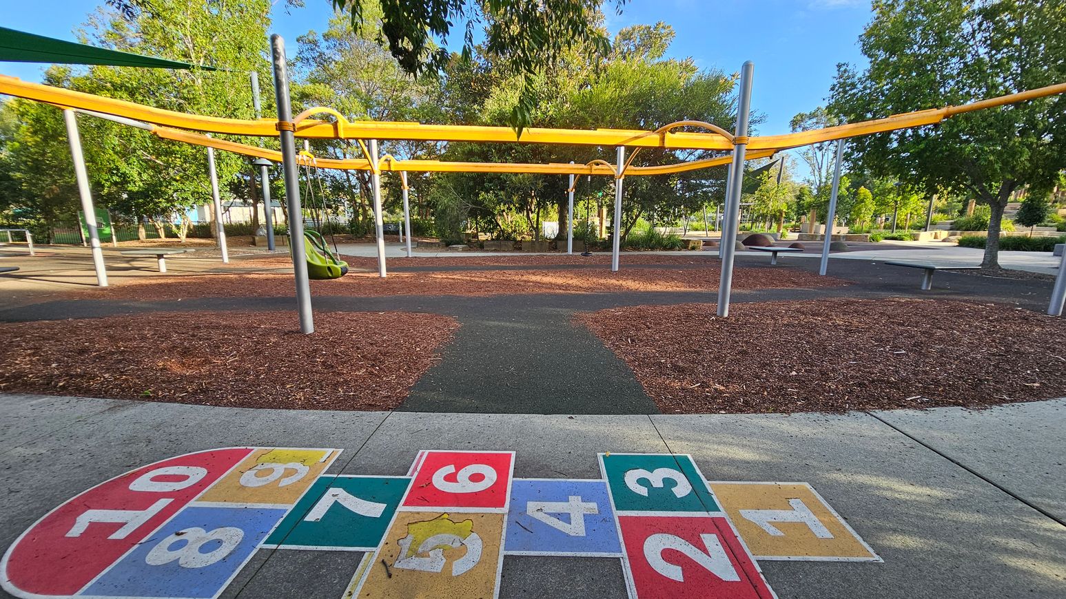 A playground with a hopscotch grid painted on the concrete in the foreground, featuring numbers 1 to 10 in colourful squares. In the middle, there are metal poles supporting yellow shade structures. A green swing hangs from one of the poles. The ground is covered with wood chips and pathways. In the background, there are trees and benches, with more greenery and a clear sky visible.