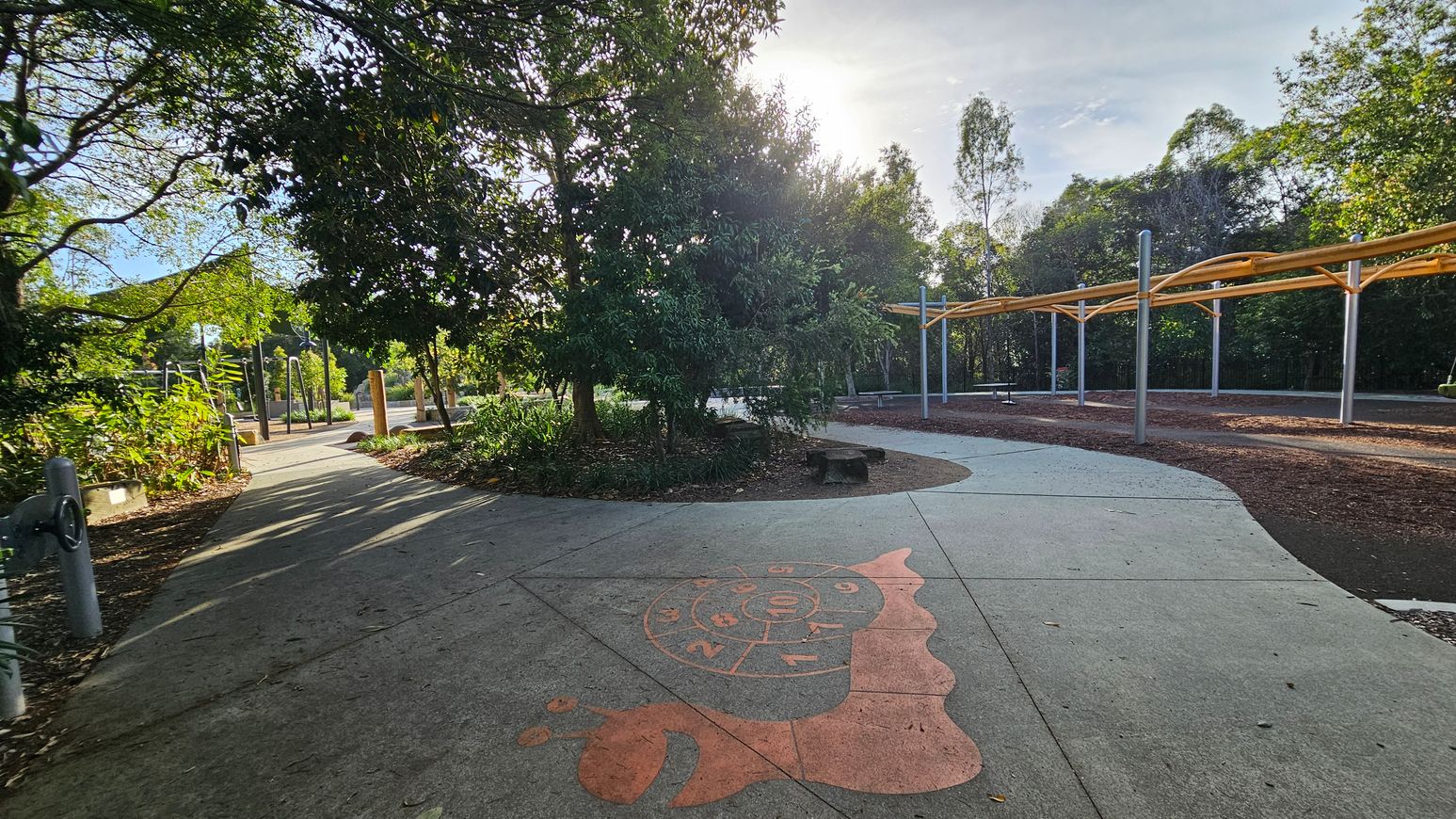 A park scene with a concrete path winding through. On the left, dense trees and foliage provide shade. In the centre, a hopscotch pattern shaped like a snail is painted on the path. To the right, a covered area with a wooden pergola supported by metal poles offers seating. The background is filled with more trees, creating a lush, green environment. The ground is a mix of concrete and mulch, and the area is well-lit by natural light.
