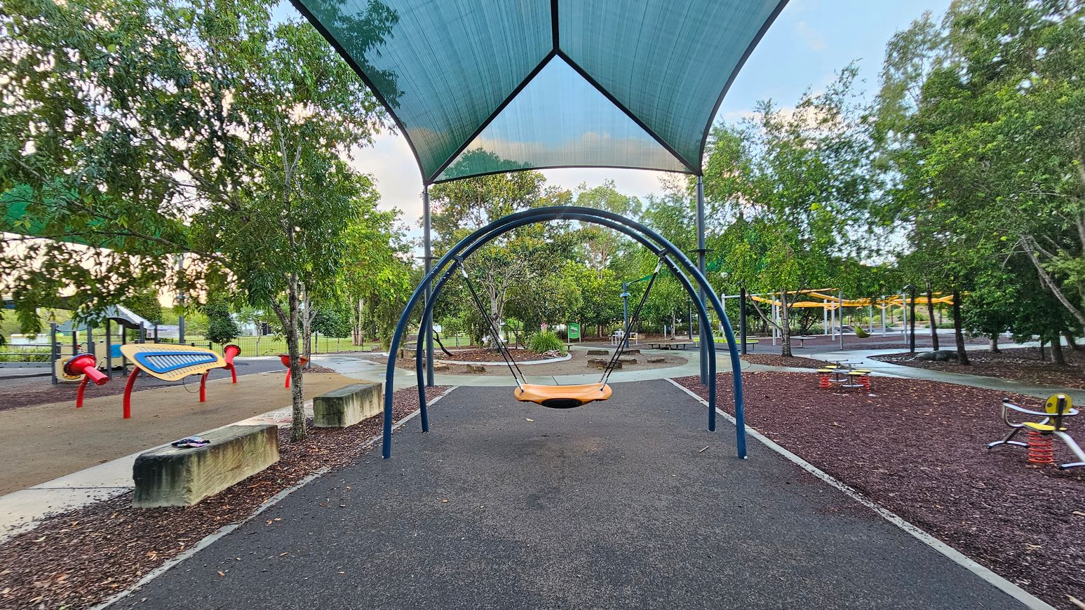 A playground with a central swing set under a large blue shade canopy. To the left, there are musical play structures with red and yellow elements. On the right, there are spring-based ride-on toys. The ground is covered with a mix of rubber and mulch. Trees surround the area, providing a green backdrop. Concrete benches are positioned near the swing, and pathways meander through the playground.