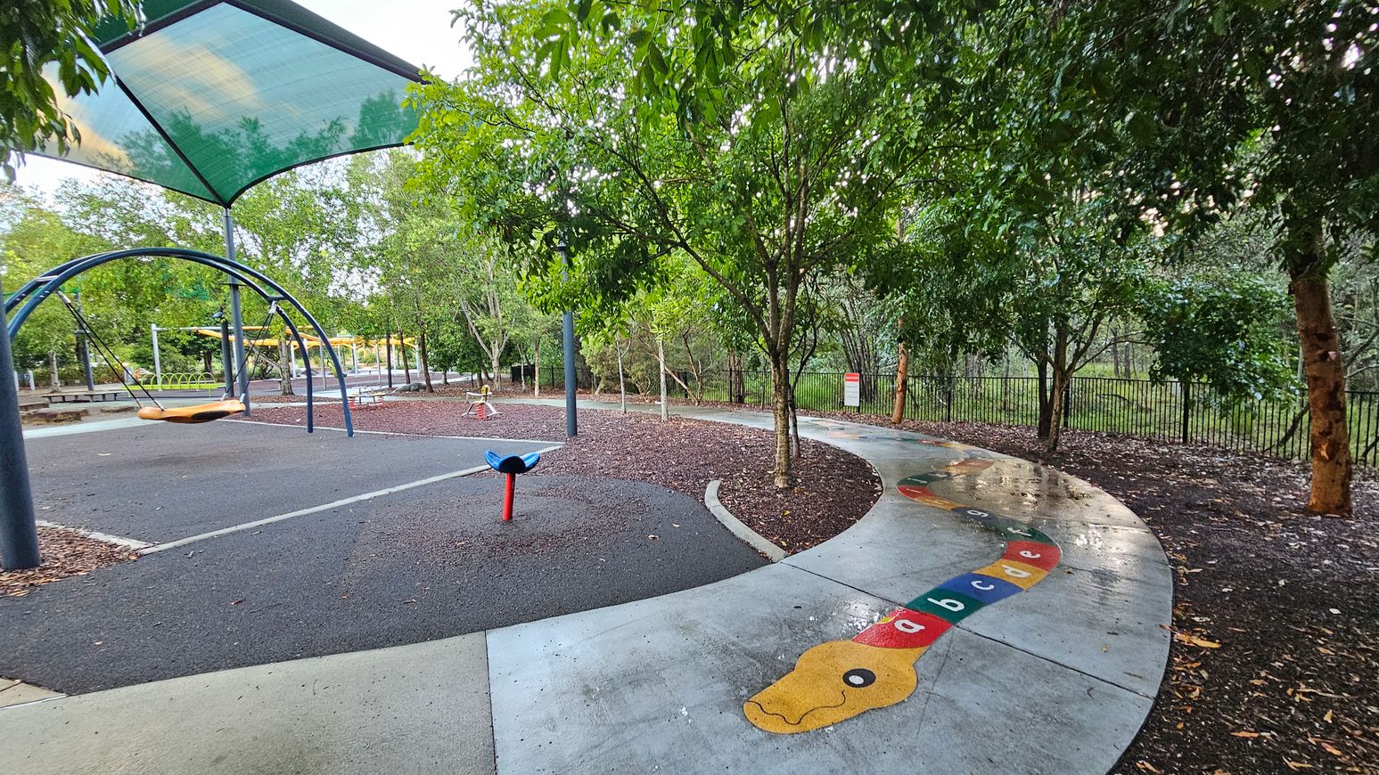 A playground with a shaded area on the left featuring a large swing set. The ground is covered with a mix of purple and grey surfaces. In the centre, a small blue and red play structure is visible. On the right, a path with a colourful snake design displaying letters of the alphabet winds through the area. Trees with lush green foliage surround the playground, and a black fence is visible in the background.