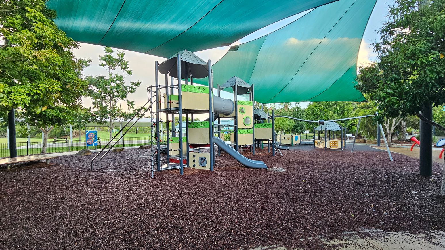 A playground with a large climbing structure in the centre, featuring slides and tunnels. The ground is covered with dark mulch. Overhead, green shade sails provide cover. Trees surround the area, with a fence and grassy field visible in the background. Benches and additional play equipment are scattered around.