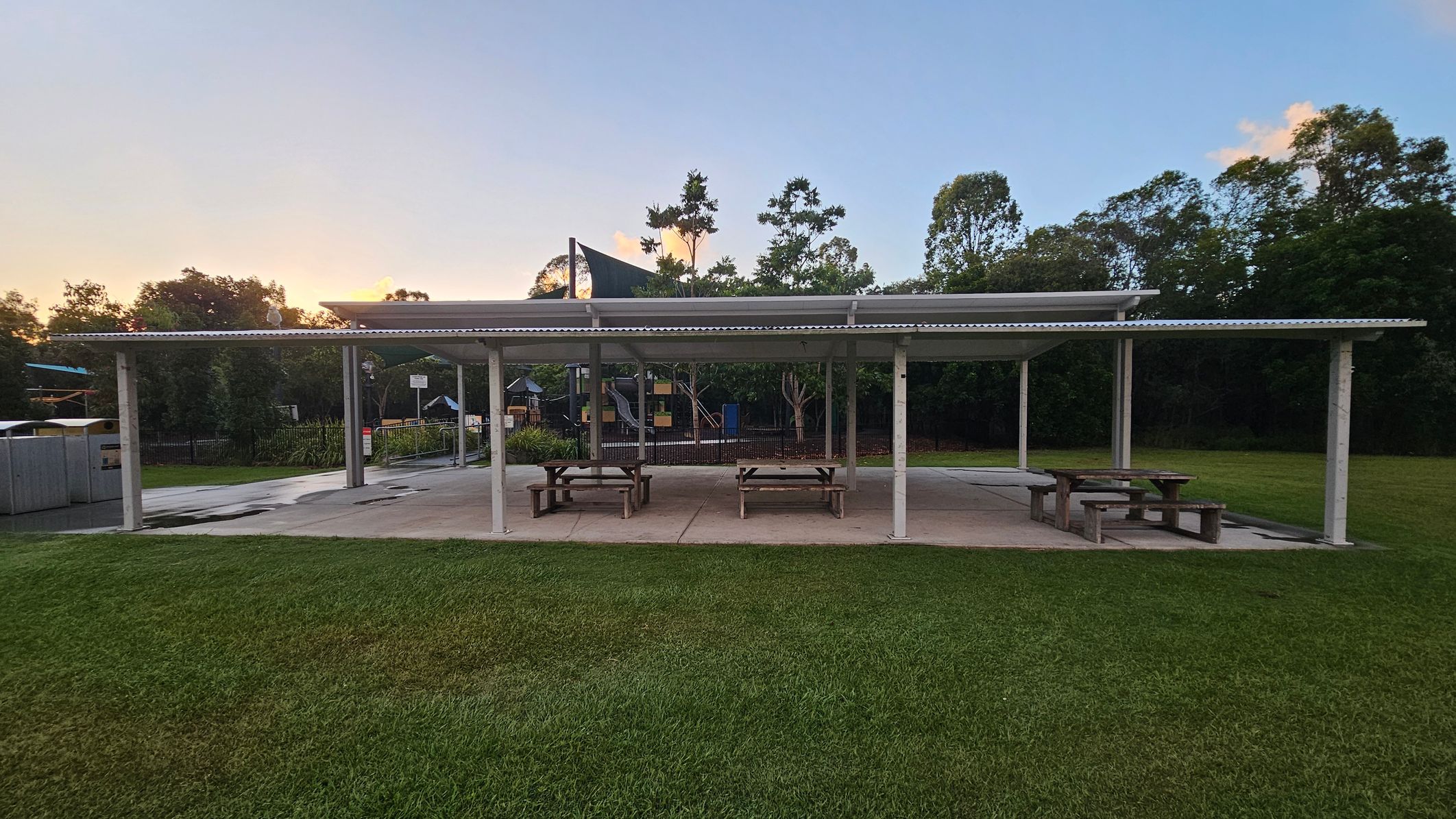 A covered outdoor area with a corrugated metal roof supported by white pillars. Beneath the shelter are several wooden picnic tables on a concrete surface. In the background, there are trees and a playground with slides and climbing structures. The foreground features a grassy lawn.